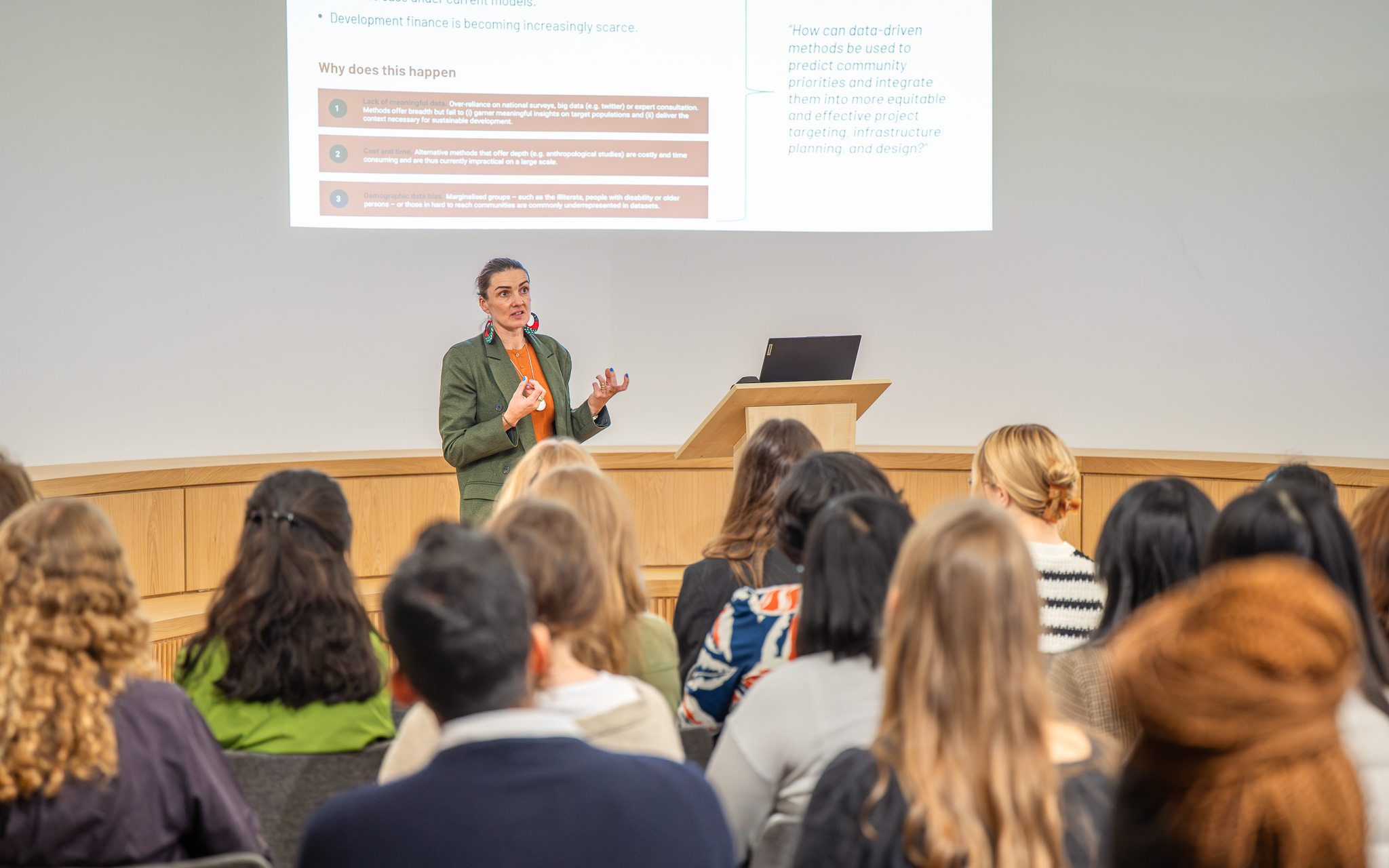 Prof Stephanie Hirmer stands at a lectern presenting to a seated audience, with a slide behind her discussing data-driven approaches to community priorities. She is speaking as part of the OxBridge Women in Engineering event.