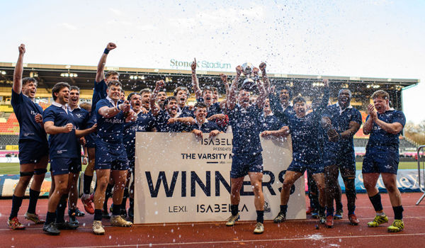 Oxford’s Men’s Blues celebrate their Varsity victory at StoneX Stadium, lifting the trophy behind a winners’ board as water sprays into the air. The team, dressed in navy kits, cheer with arms raised on the pitch following their win over Cambridge.